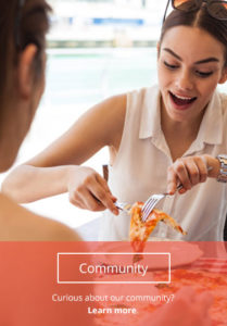 A woman at a restaurant smiles while serving herself a slice of pizza with a man across the table from her. A button on the bottom of the image reads, "Community."